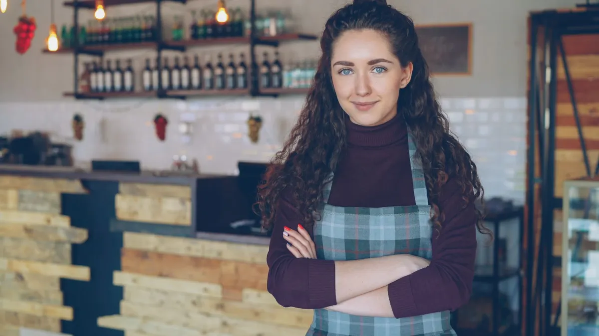 apron, bar, bartender, bottles, business, commercial, company, corporate, counter, curly, curly hair, enterprise, female, hair, office, owner, portrait, professional, rustic, shop owner, small, small business, smiling, standing, wooden, young